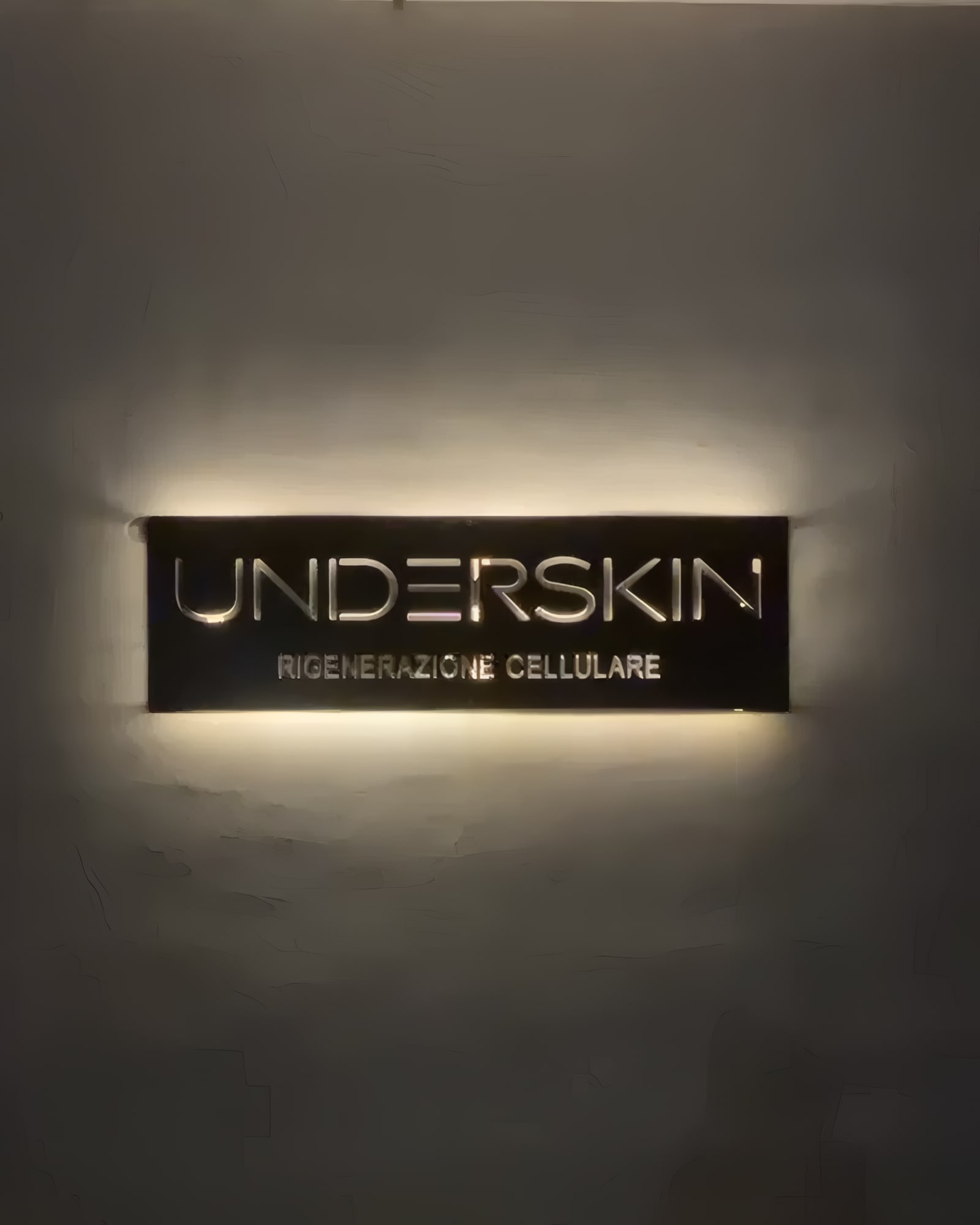 Insegna Underskin retroilluminata di sera: la luce calda filtra dalle lettere traforate e disegna un alone sulla parete