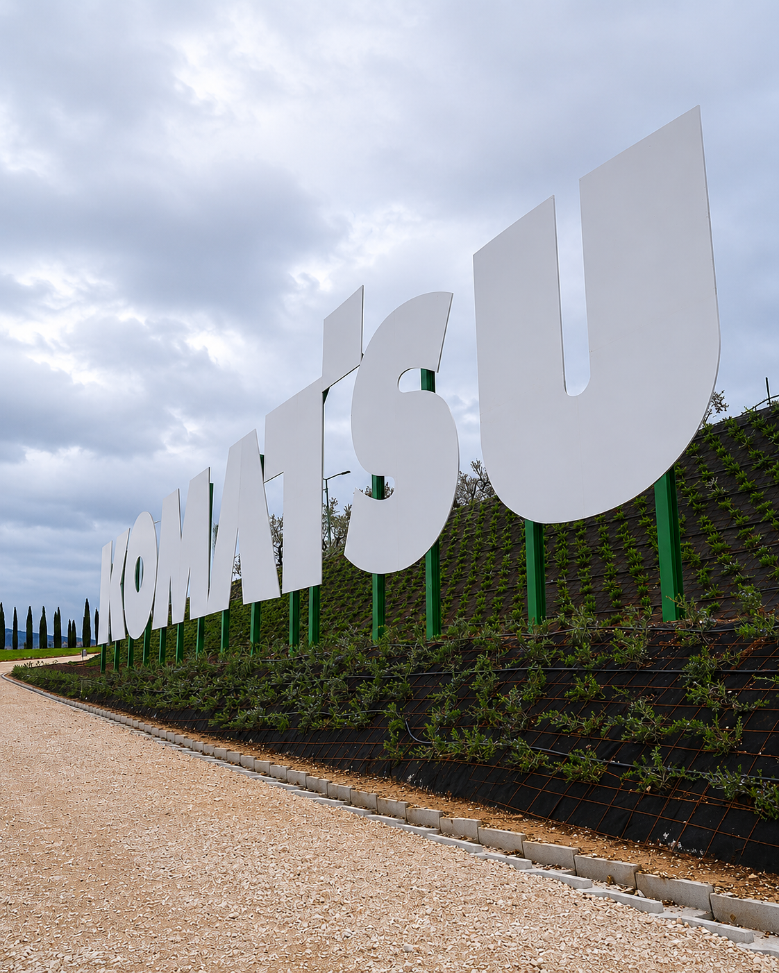 Installazione monumentale con le lettere KOMATSU in acciaio realizzata da Æstus Design per GIS Group, landmark d'ingresso integrato nella collina verde